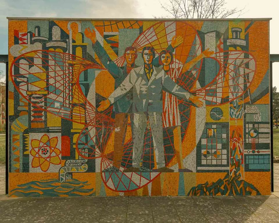 On one side, there is the Makrokosmos with two cosmonauts venturing into the vast expanse of space, reflecting the Soviet pride in the Space Race. On the other side, three scientists stand before symbols of atomic energy, illustrating the “promise” of nuclear power for a modern society.
