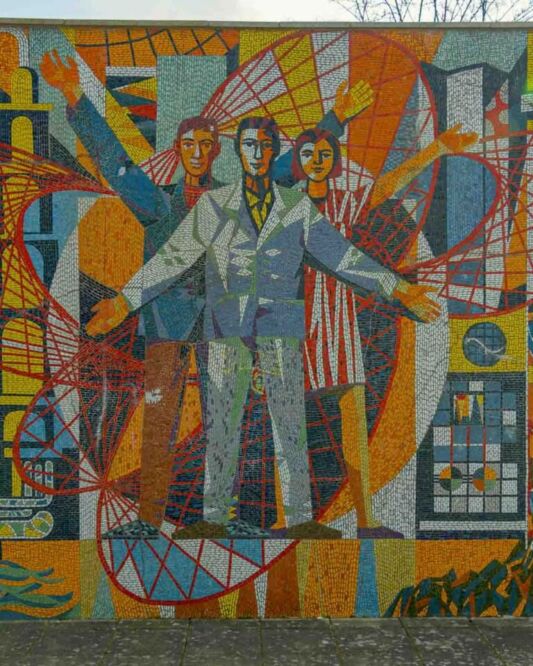 On one side, there is the Makrokosmos with two cosmonauts venturing into the vast expanse of space, reflecting the Soviet pride in the Space Race. On the other side, three scientists stand before symbols of atomic energy, illustrating the “promise” of nuclear power for a modern society.