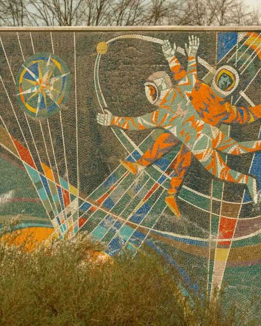 On one side, there is the Makrokosmos with two cosmonauts venturing into the vast expanse of space, reflecting the Soviet pride in the Space Race. On the other side, three scientists stand before symbols of atomic energy, illustrating the “promise” of nuclear power for a modern society.